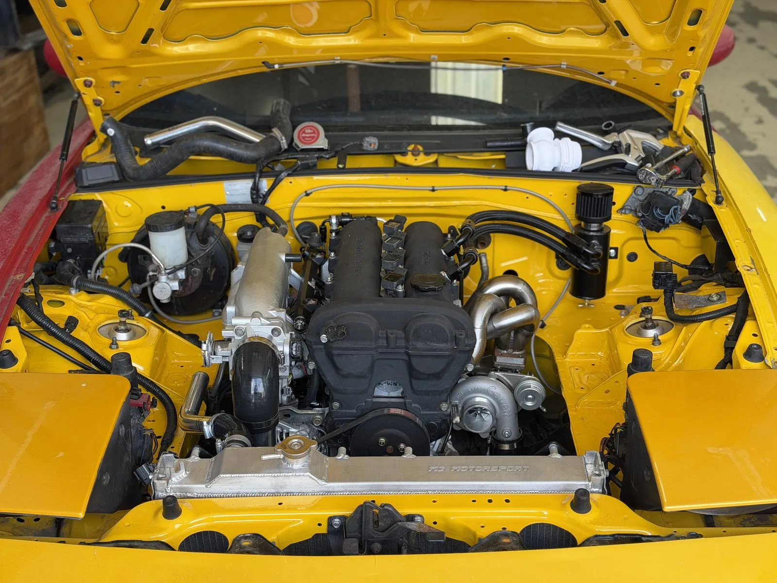 A black baffled rocker cover fitted in a yellow turbocharged MX-5 engine bay