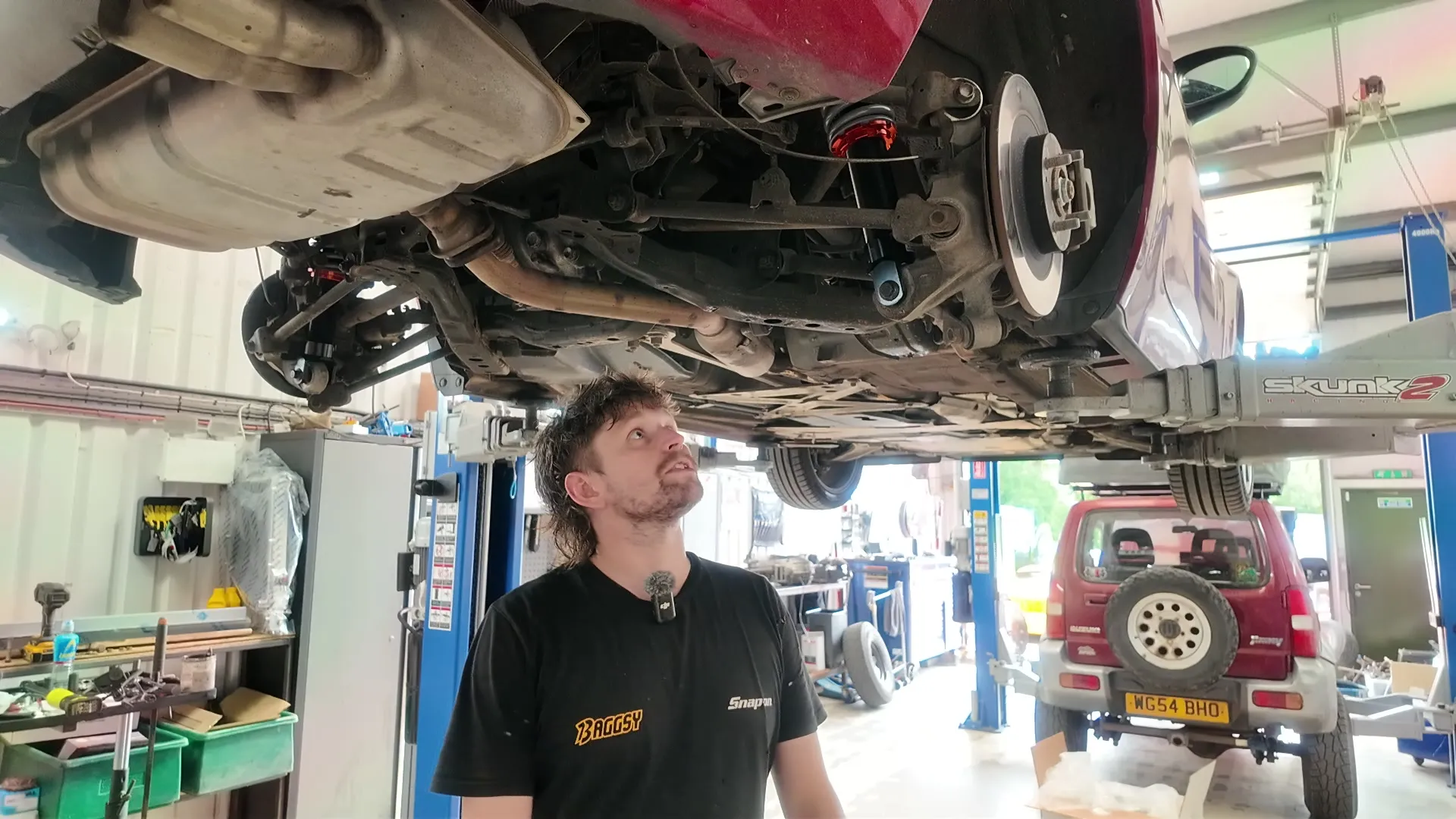 Under the car on the lift, explaining the bushing preload reset with the rear suspension overhead