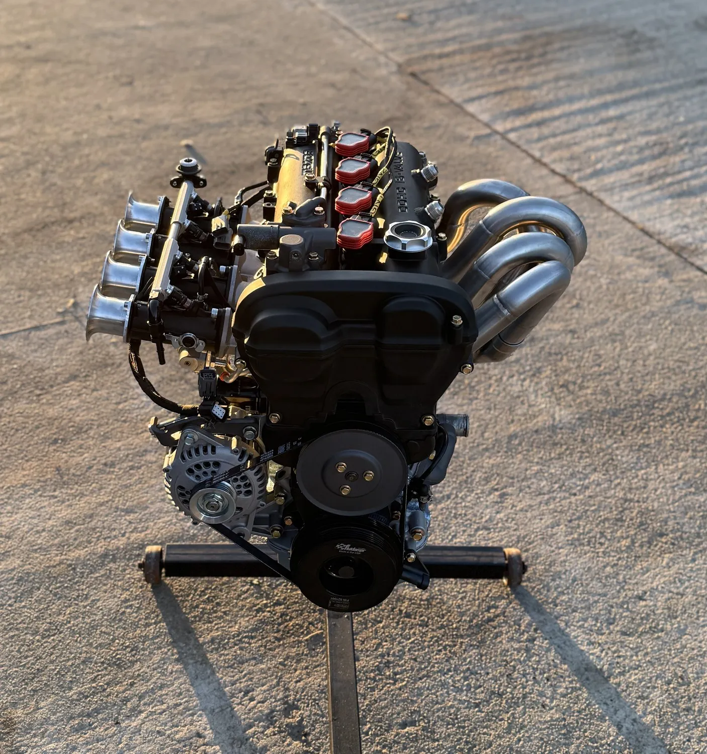The dressed engine on its stand outside with the FOJI Raceworks exhaust manifold fitted, tubes catching the late sun