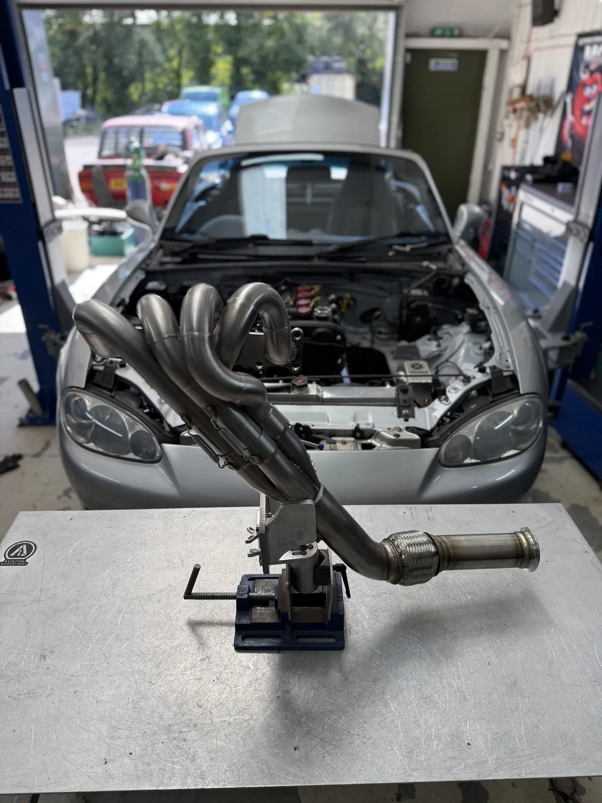 The FOJI Raceworks high rise manifold and downpipe clamped in a vice, with the Mk2.5 it was built for behind it on the ramp