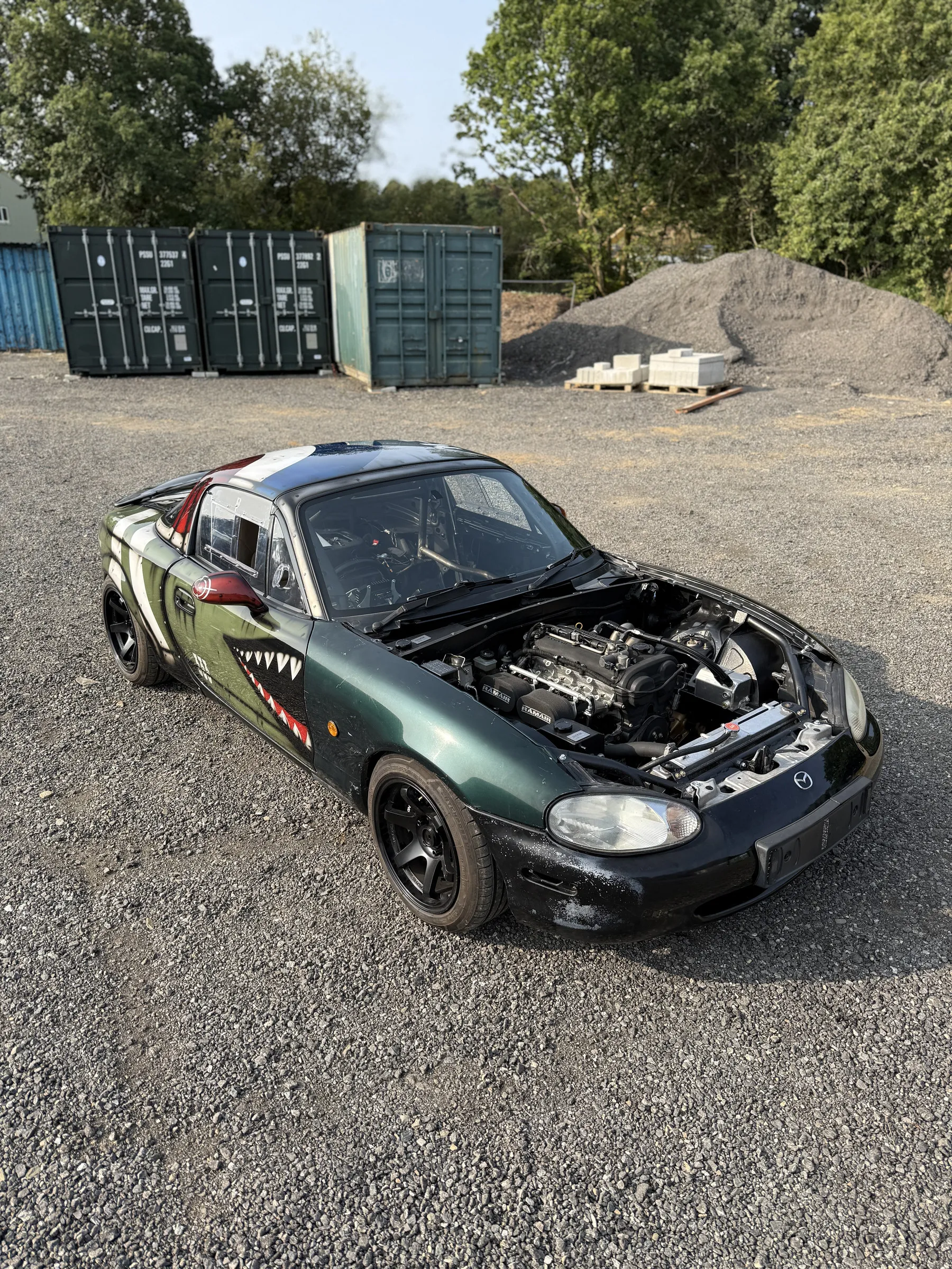 Moxie outside the workshop on the gravel with the bonnet up, shipping containers behind