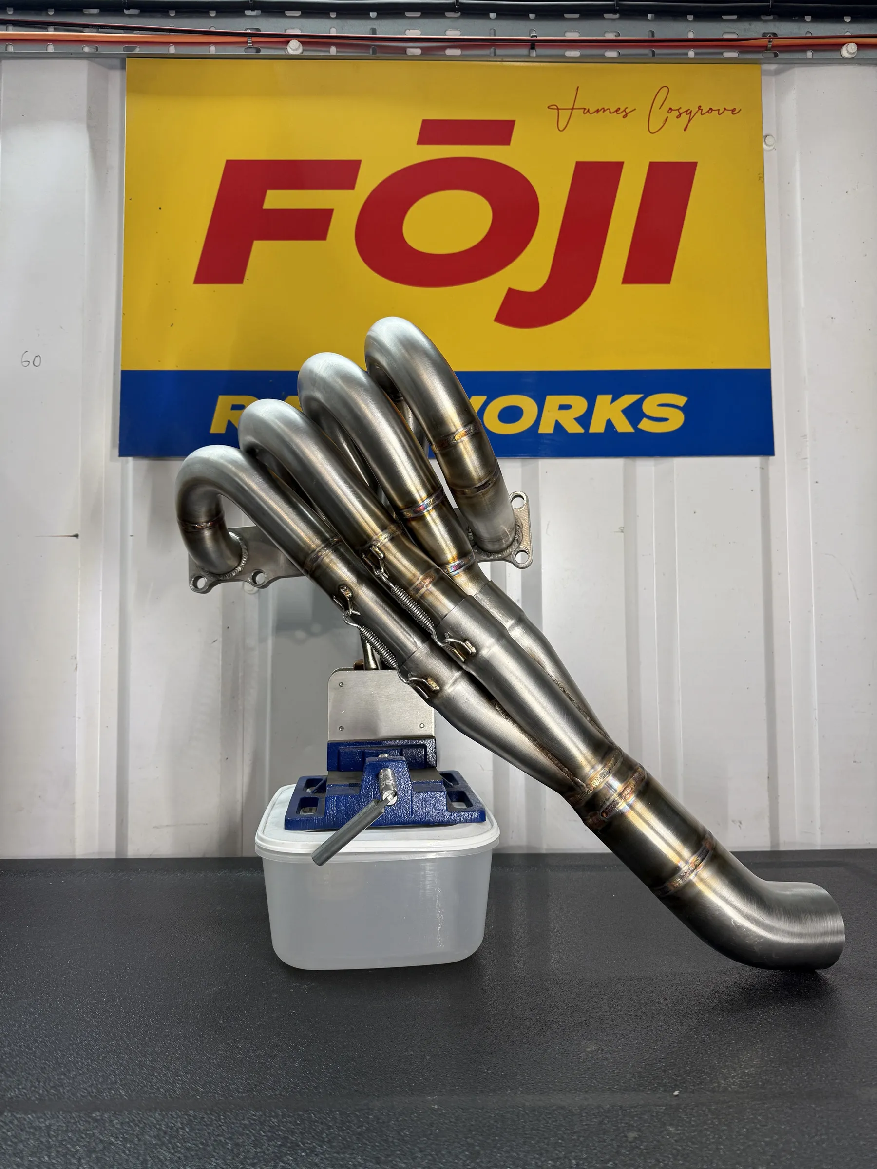 The finished four into one tubular manifold clamped on the bench in front of a FOJI Raceworks banner, heat tinted runners sweeping into the collector