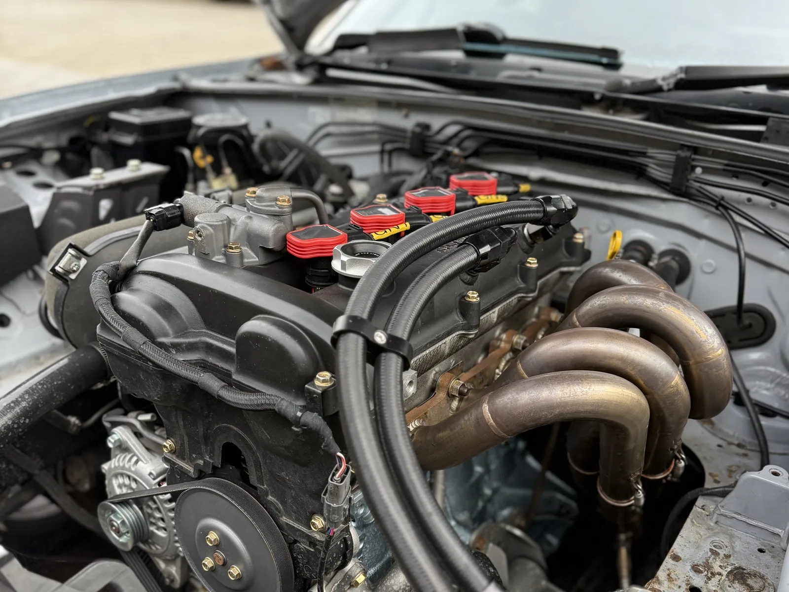 A finished rocker cover fitted in an MX-5 engine bay, braided breather lines running off it beside the exhaust manifold