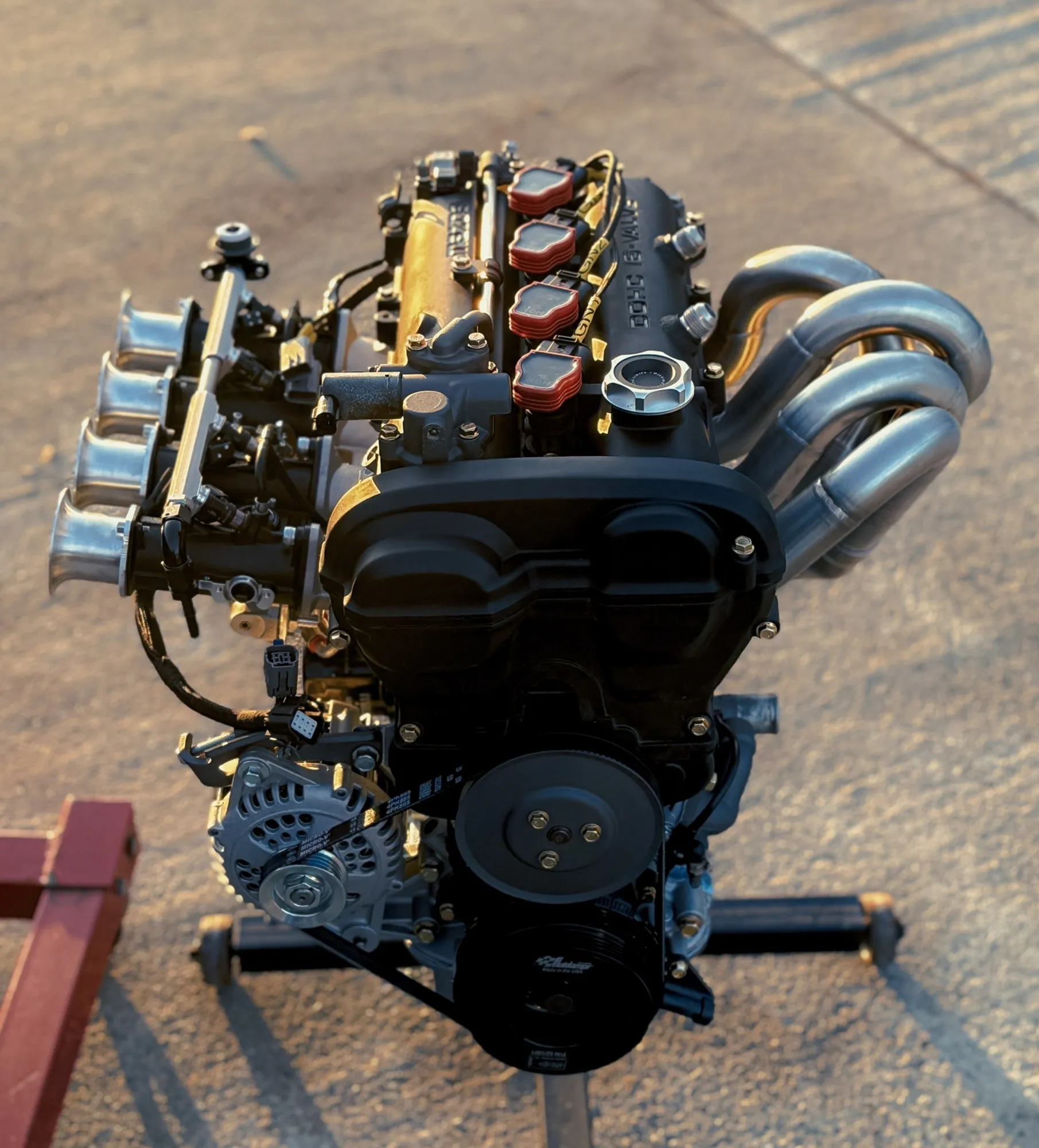 A finished black rocker cover on a built MX-5 engine on its stand in the late sun, individual throttle bodies on one side and a tubular manifold on the other